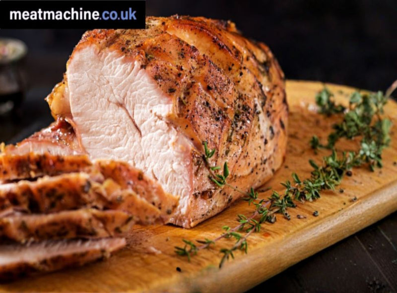 Sliced roasted pork on a wooden board with herbs, branded 'meatmachine.co.uk'.