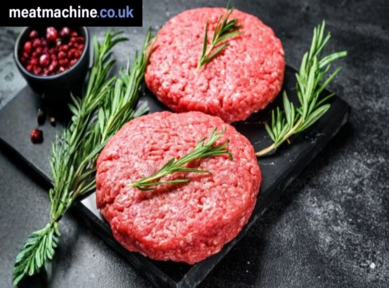 Three raw burger patties on a black slate board with rosemary sprigs, accompanied by a small bowl of spices, on a dark surface.